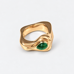 A gold ring with an organic, wavy shape and a green oval gemstone, photographed on a light gray background.