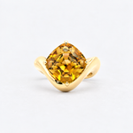 A yellow gold ring featuring a cushion-cut yellow gemstone in a prong setting, displayed on a clean light gray background.