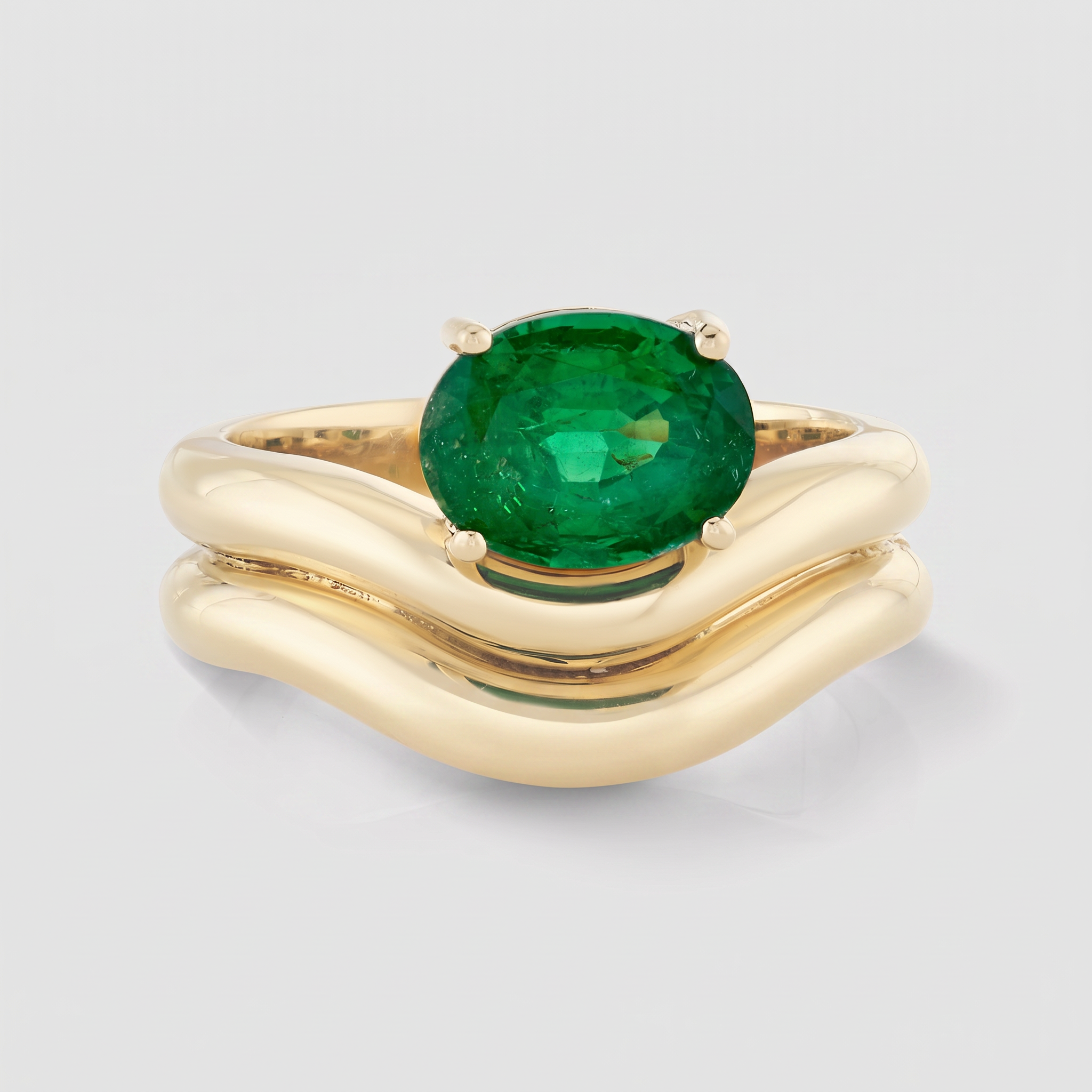 A yellow gold ring with a wavy, layered band and a large oval-cut green emerald, shown on a light gray background.