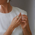 A close-up lifestyle shot of a woman wearing a textured silver ring on her finger and a matching white gold baguette earring, dressed in a white top against a neutral gray background.