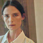 A close-up portrait of a woman with dark hair pulled back, wearing a white collared shirt and long green beaded drop earrings against a warm brown and beige background.