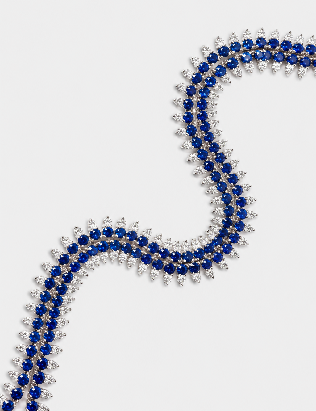 A close-up of a double-row necklace featuring deep blue sapphires and diamonds in a silver-toned setting, arranged in a curved S-shape on a light gray background.