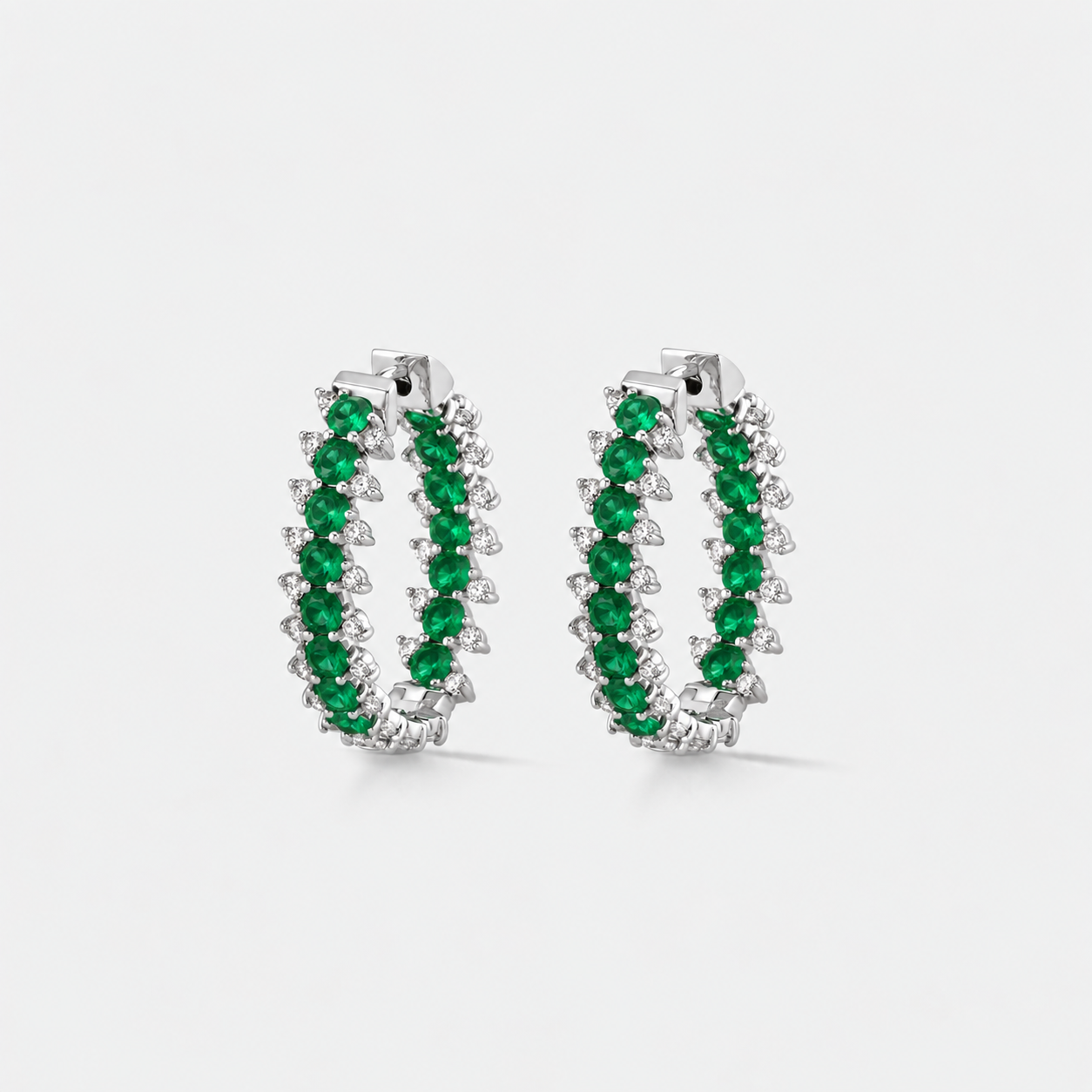 A pair of silver hoop earrings embellished with rows of round green emeralds and small diamonds, shown on a light gray background.