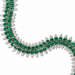 A close-up of a luxury necklace featuring rows of round-cut emerald green gemstones and pear-cut clear gemstones in a white-toned setting, arranged in a curved S-shape on a solid white background.