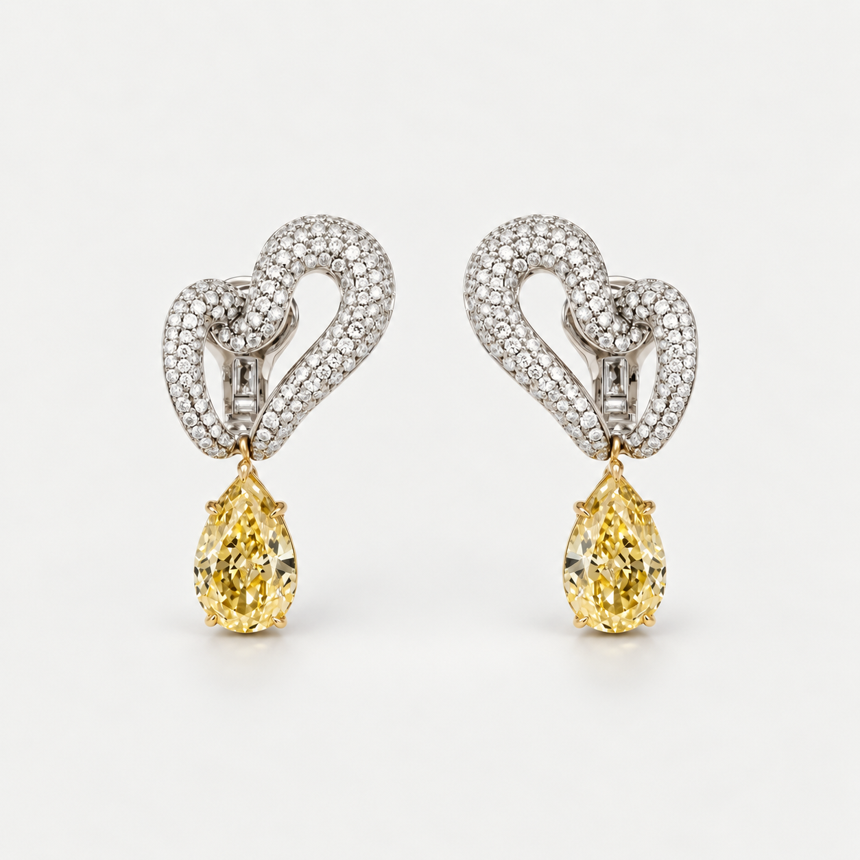 A pair of high-end diamond earrings featuring a pavé heart design in white metal with a dangling pear-cut yellow gemstone below, set against a clean white background.
