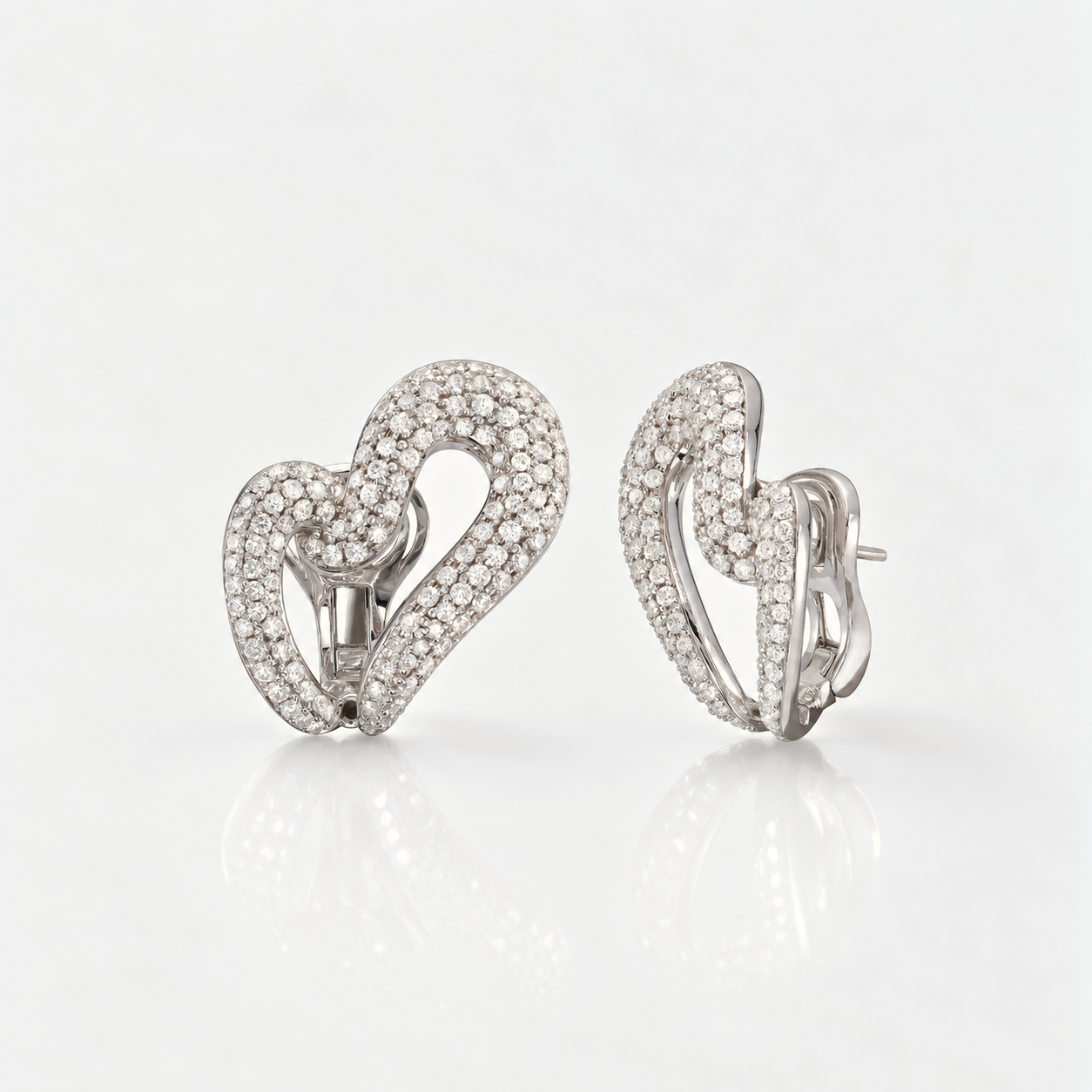 A pair of silver-toned heart-shaped earrings paved with small clear stones, shown from the front and side on a clean white reflective surface.