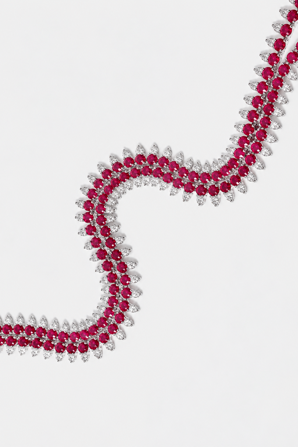A close-up of a silver-toned necklace featuring rows of round deep pinkish-red rubies and round diamonds, arranged in a flowing S-shape on a light gray background.