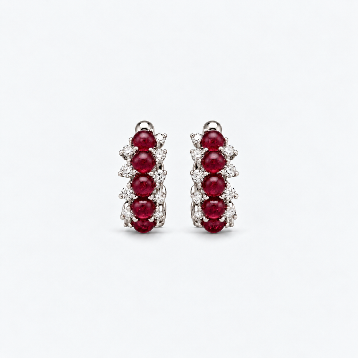 A pair of silver-toned huggie hoop earrings featuring three round-cut deep red rubies and small clear stones, set against a clean white background.