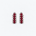 A pair of silver-toned huggie hoop earrings featuring three round-cut deep red rubies and small clear stones, set against a clean white background.