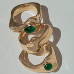 Three interlocking gold rings with oval green gemstone accents, arranged diagonally on a light gray background.