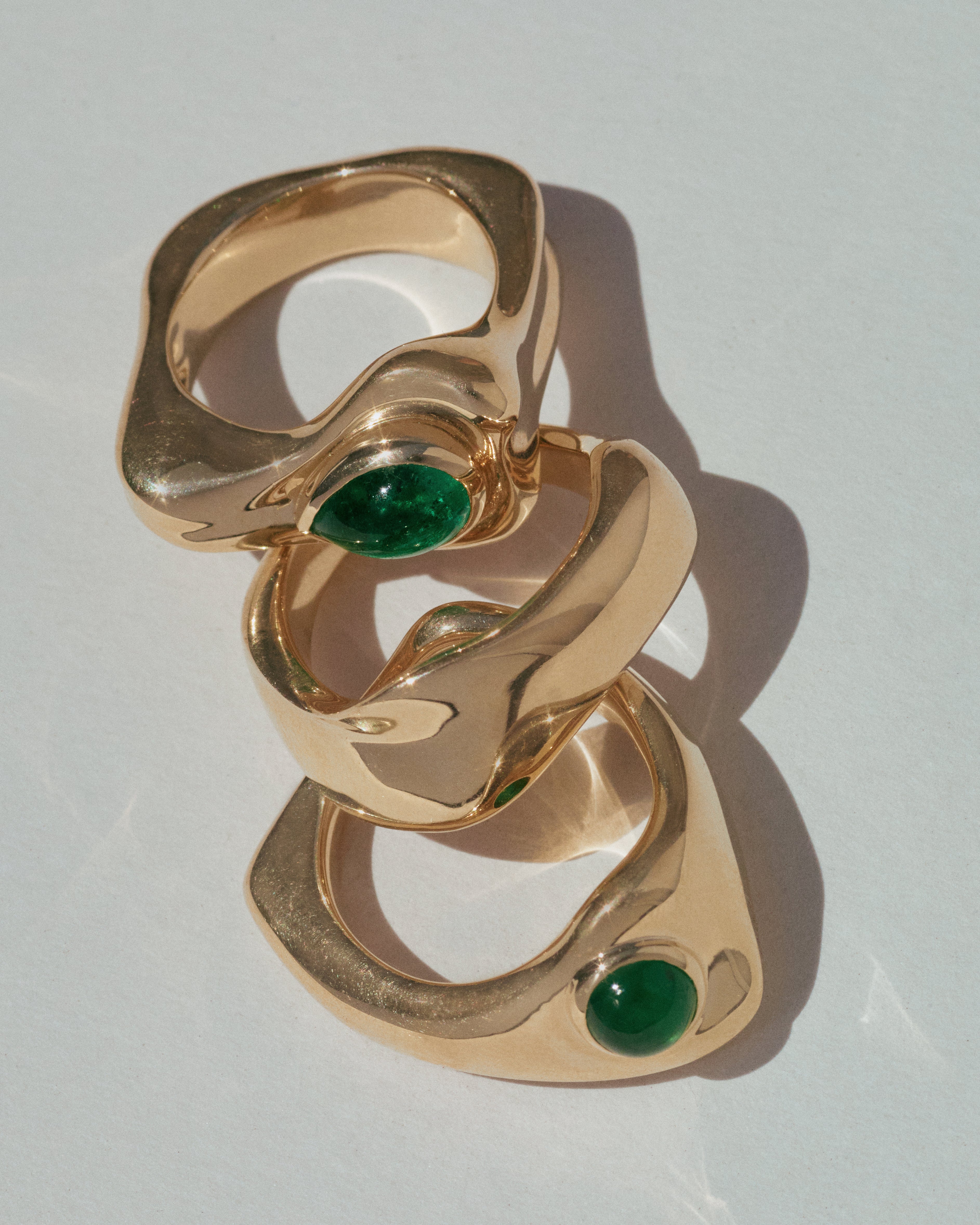 Three interlocking gold rings with oval green gemstone accents, arranged diagonally on a light gray background.